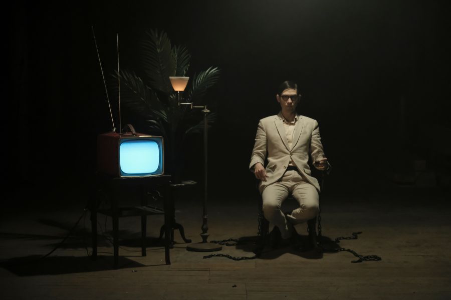 Still image from Black Lodge's film featuring Timur sitting in a chair in a dimly lit room, next to a TV