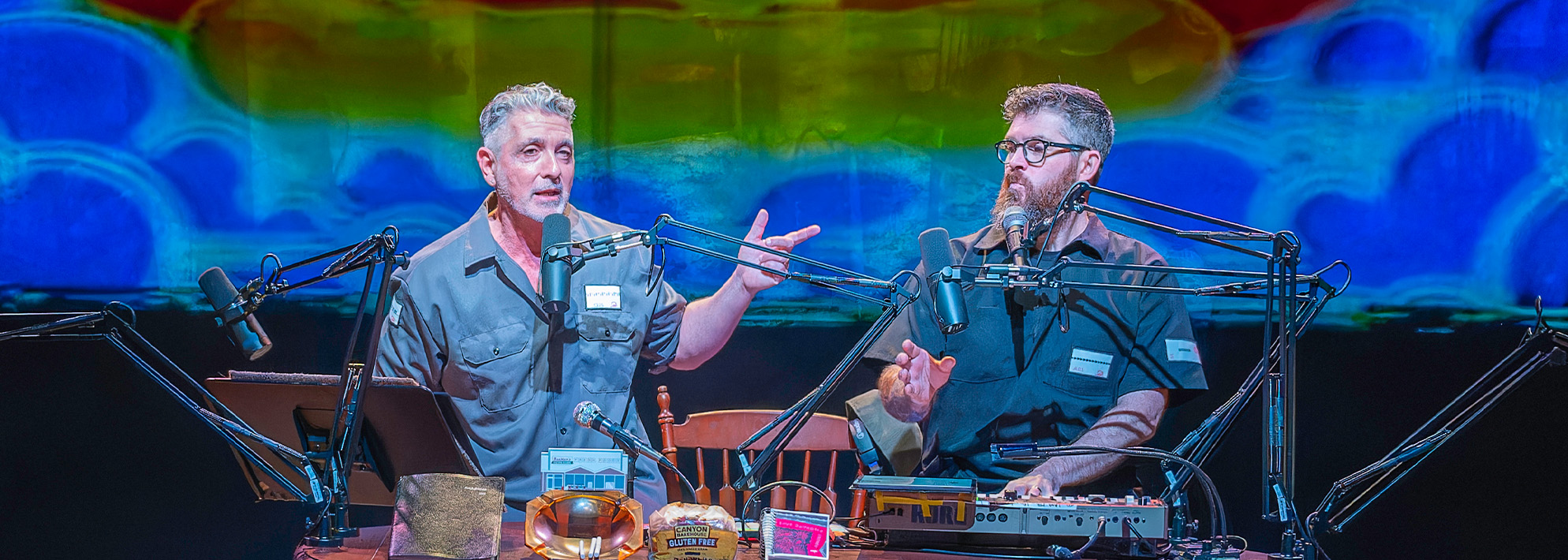 Ain Gordon and Josh Quillen sit at a table on stage with a projection behind them of a painting of a rainbow