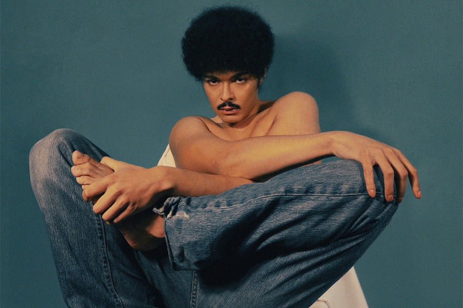 Image of Isaac Dunbar posed in a chair with legs crossed in front of blue backdrop