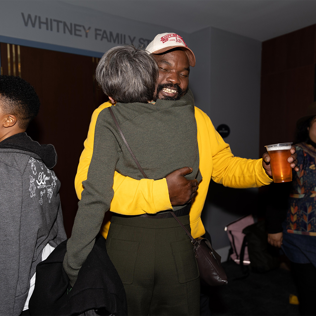 Two people embrace in the lobby of the The Nimoy. One wears a grey sweater and is faced away from the camera. The other wears a yellow sweatshirt and a white baseball cap, holds a glass, and grins widely as they embrace.