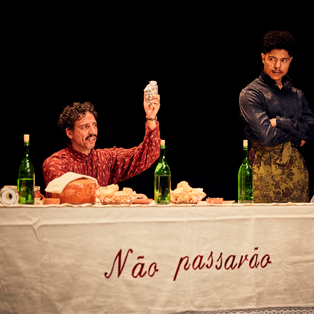 A still from 'Catarina' featuring two actors. One sits at a table with food and three wine bottles before him and holds up a silver item as he speaks. A man next to him stands with his arms crossed, listening on with a serious expression.