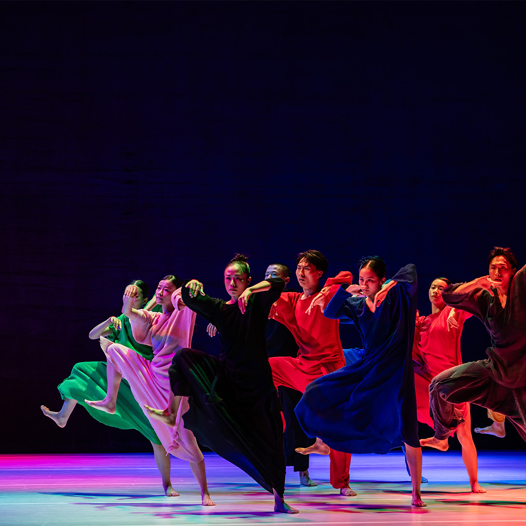 Members of Cloud Gate march across a rainbow lit stage wearing colorful gowns. They move in unison with one leg raised, mid large step.