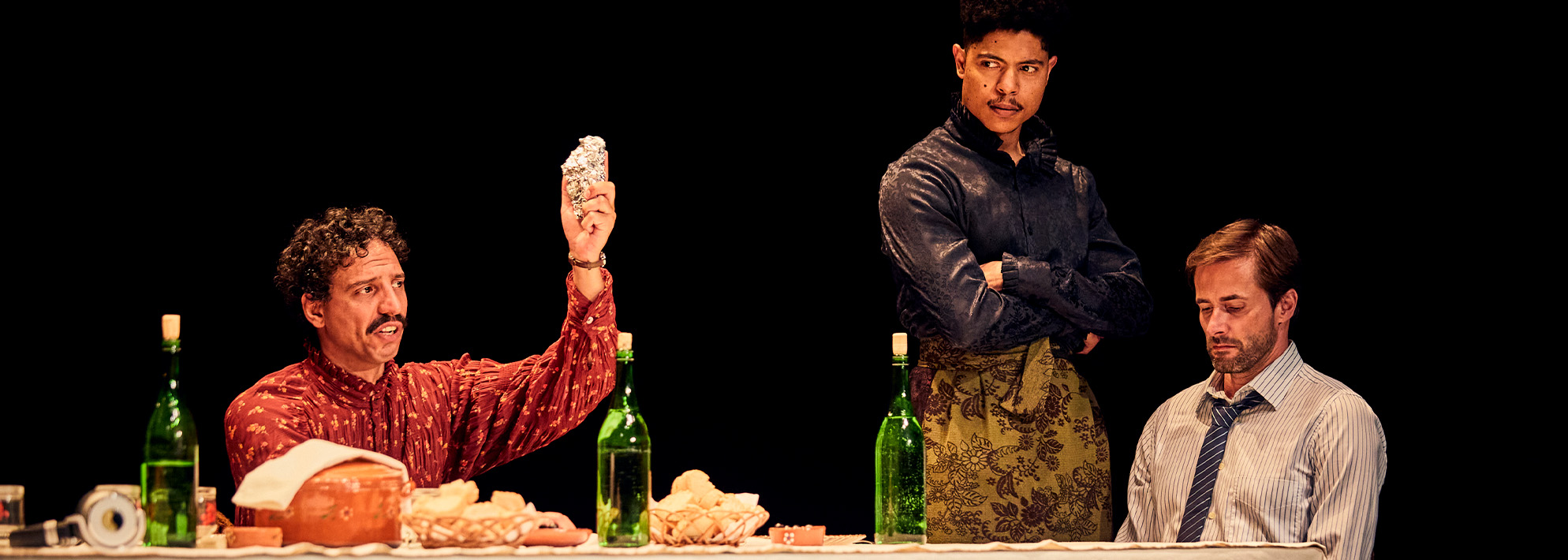 A still from 'Catarina' featuring two actors. One sits at a table with food and three wine bottles before him and holds up a silver item as he speaks. A man next to him stands with his arms crossed, listening on with a serious expression.