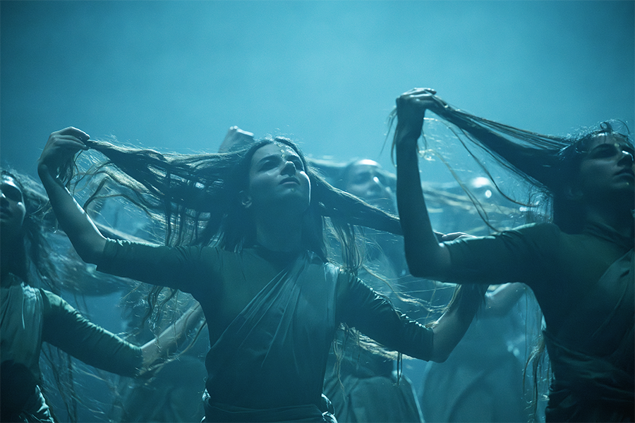 Dancers pose in blue light on stage. They grab onto pieces of their own hair and hold it outstretched at the side of their head.