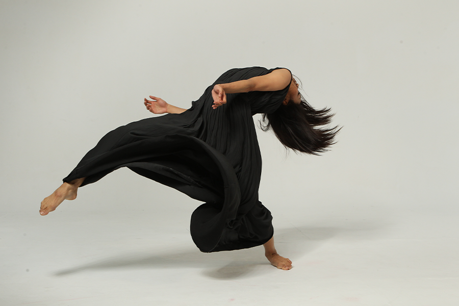 A dancer poses in front of a white backdrop while wearing loose fitting all black clothing.  The dancer leans backwards, their head obscured by their hear which is undone and moving with the motion of the pose.