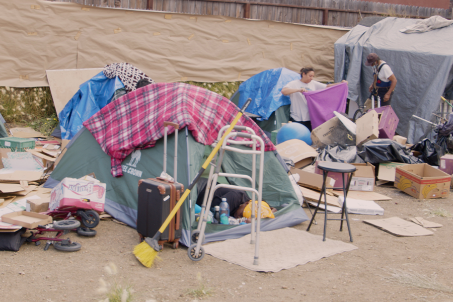 Still from Crushing Wheelchairs featuring image of homeless encampment community featuring a green tent at the center. 2 people tend to the encampment, organizing items from it.
