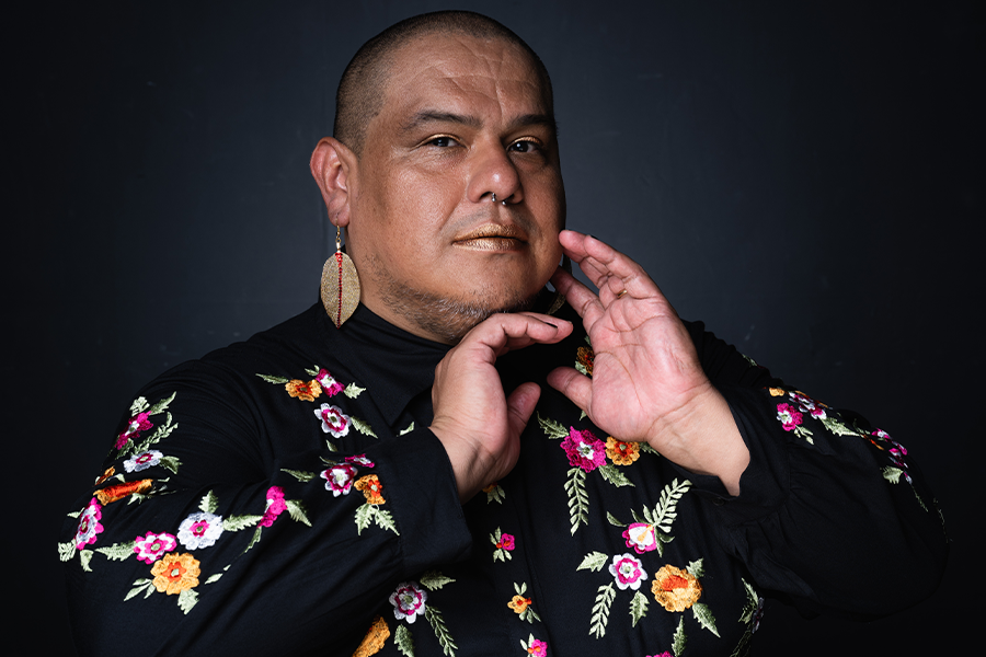 Dorian Wood poses in a floral embroidered black gown in front of a black backdrop. They have one hand positioned underneath their chin and the other lightly touching their face.