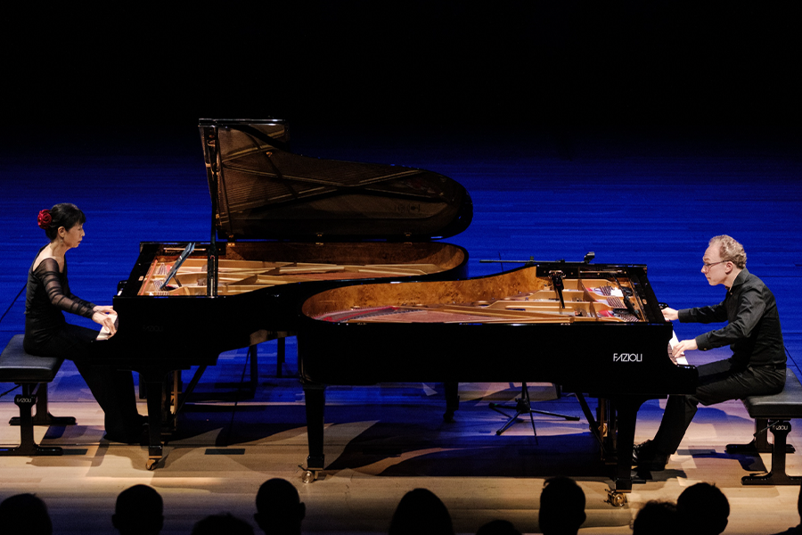 Gloria Cheng and Ralph van Raat perform on stage, lit by blue lighting. Both artists perform at their own pianos, positioned to be facing one another. The silhouettes of some audience members are in the foreground.