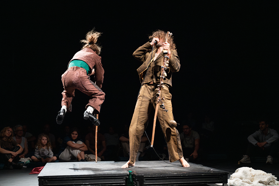 Performance image from Mirlitons featuring Hainaux and Chaignaud performing in the center of a theater on a small square stage. Silhouettes of audience members are seen in the foreground and background. Chaignaud faces away from the camera as he lifts on leg in the air, tap dancing. He wears pink pants and a jacket, holding a large wooden stick. Hainaux faces towards the camera and Chaignaud, wearing a brown suit and holding a wooden stick with bells attached to it. 
