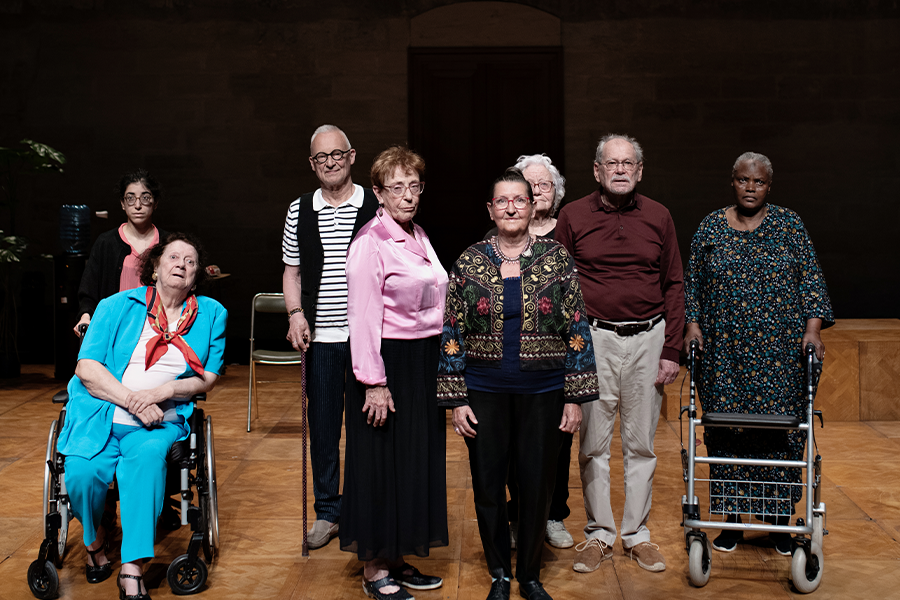 The cast La Vie Secrète des Vieux, 8 people in total, poses together on stage, staring directly into the camera. The floor underneath them is a light wood.