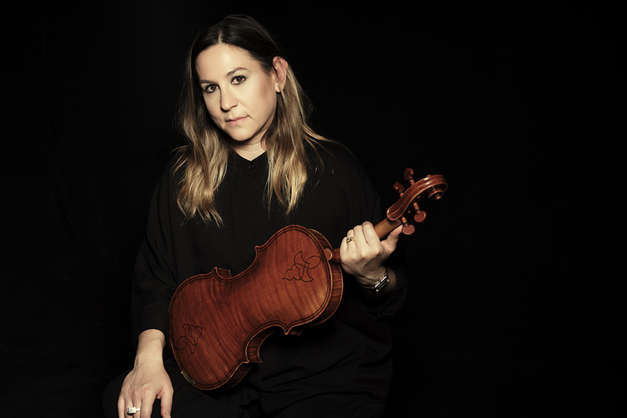 Nadia Sirota poses in front of a black backdrop wearing a black blouse, holding a viola as they look direct to camera