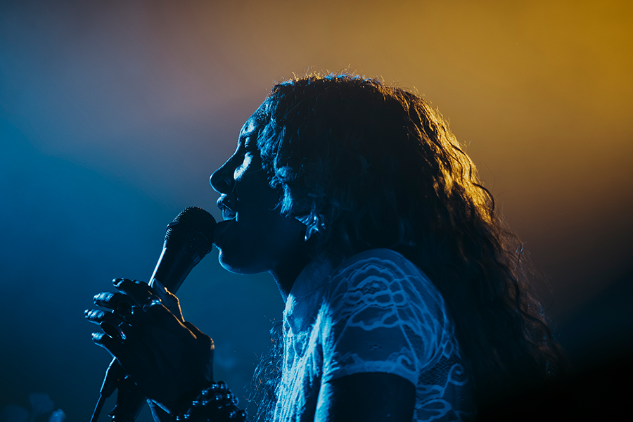 Side profile photo of SPELLLING performing on stage wearing a white top. SPELLLING sings into a microphone in this close up show, illuminated by blue and yellow lighting.