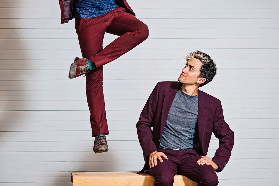 Caleb Teicher sits on a wooden bench wearing a maroon suit and a blue tee shirt. They look up as Nic Gareiss, wearing burgundy trousers, jumps up in the air with one leg crossed over the other, only the lower portion of their body/legs in frame.