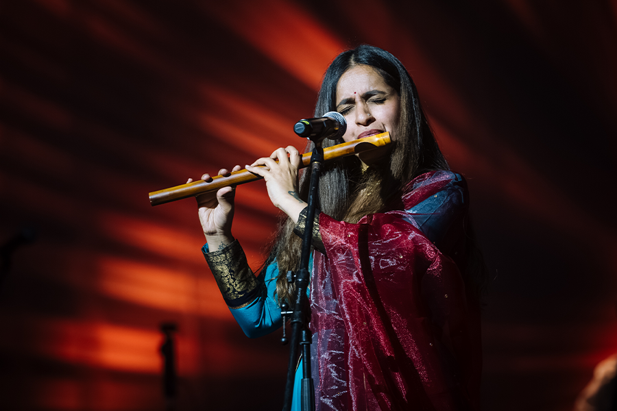 Varijashree Venugopal performs on stage with orange rays of light in the background behind her. Venugopal plays the flute into a microphone while wearing colorful purple and blue garments.