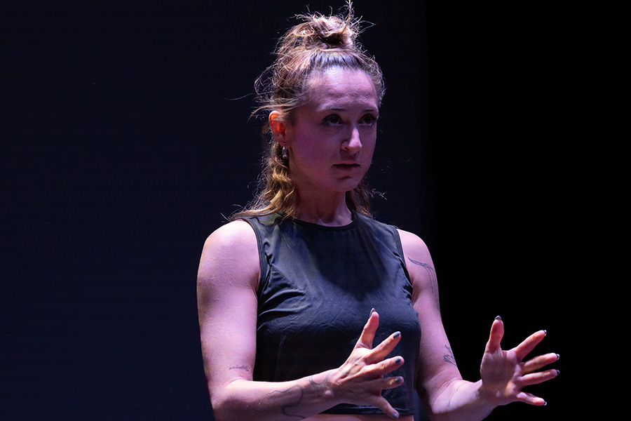 Image of a dancer on stage, illuminated by purple tinged lighting. The dancer wears their hair in a bun, a black tank top, and holds their hands outwards in front of them while seeming to be addressing someone off camera.