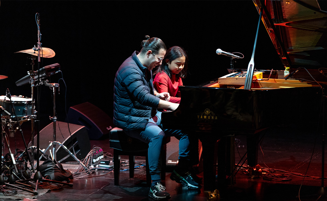 Alfredo Rodriguez performs on stage with a student at a DFS performance. The two play the piano.