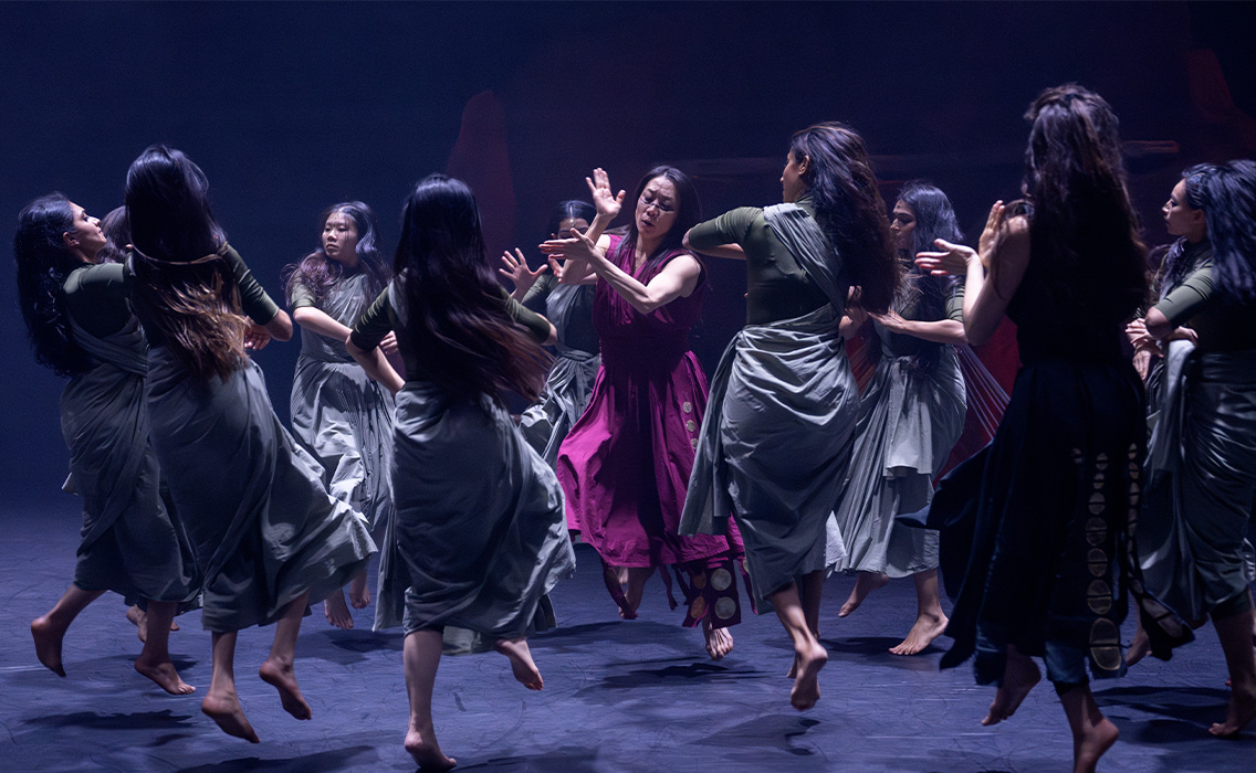 Image from Akram Khan's 'Thikra,' featuring dancers wearing dresses ad performing on stage, moving towards each other in a center