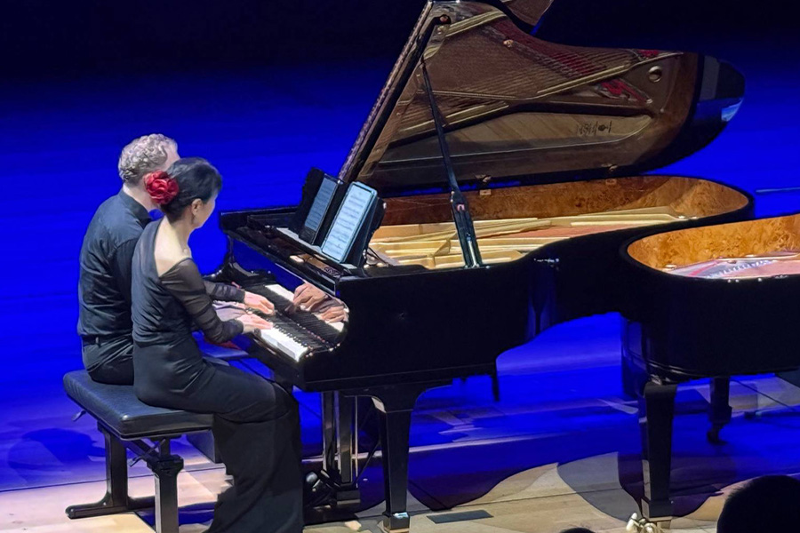 Gloria Cheng and Ralph van Raat perform on stage, lit by blue lighting. Both artists perform at their own pianos, positioned to be facing one another. The photo is taken from a high angle, showing the silhouettes of some audience members in the foreground.
