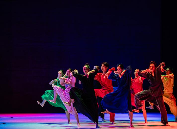 Members of Cloud Gate march across a rainbow lit stage wearing colorful gowns. They move in unison with one leg raised, mid large step.
