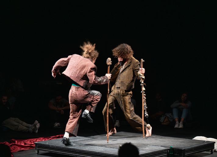 Performance image from Mirlitons featuring Hainaux and Chaignaud performing in the center of a theater on a small square stage. Silhouettes of audience members are seen in the foreground and background. Chaignaud faces away from the camera as he lifts on leg in the air, tap dancing. He wears pink pants and a jacket, holding a large wooden stick. Hainaux faces towards the camera and Chaignaud, wearing a brown suit and holding a wooden stick with bells attached to it. He beatboxes into a microphone.