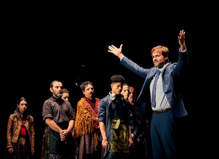 A performer on stage poses with outstretched arms and cast members in the foreground. 
