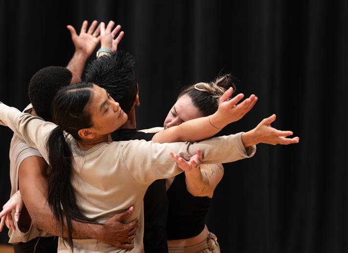Four contemporary dancers embrace during a rehearsal, creating a sculptural moment of movement, connection and collaboration in a performing arts studio.