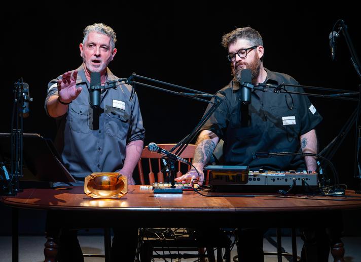 Two male performers sit at a podcast-style table with microphones, sound equipment, and props, delivering a live multimedia performance in front of a colorful, abstract projection