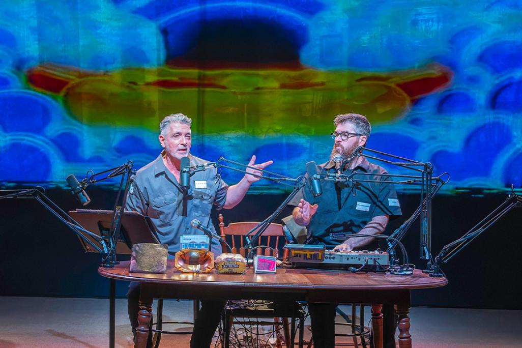 Two male performers sit at a podcast-style table with microphones, sound equipment, and props, delivering a live multimedia performance in front of a colorful, abstract projection