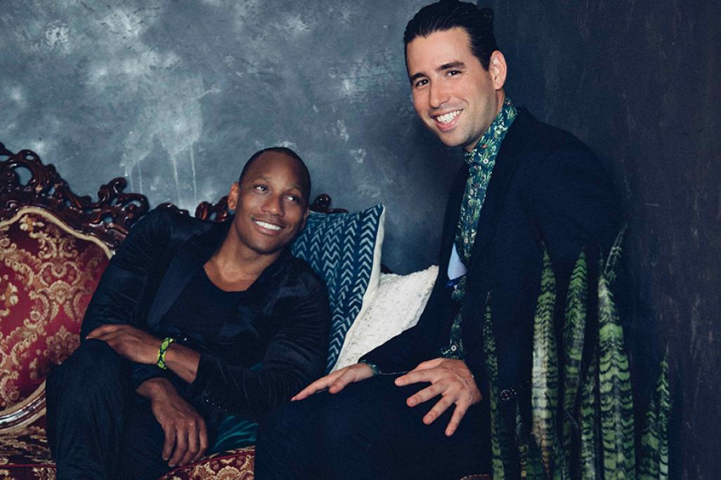 Alfredo Rodriguez and Pedrito Martinez relax on a vintage red-upholstered couch against a textured gray wall, exuding warmth and charm in a cozy, stylish setting accented by patterned pillows and a leafy plant
