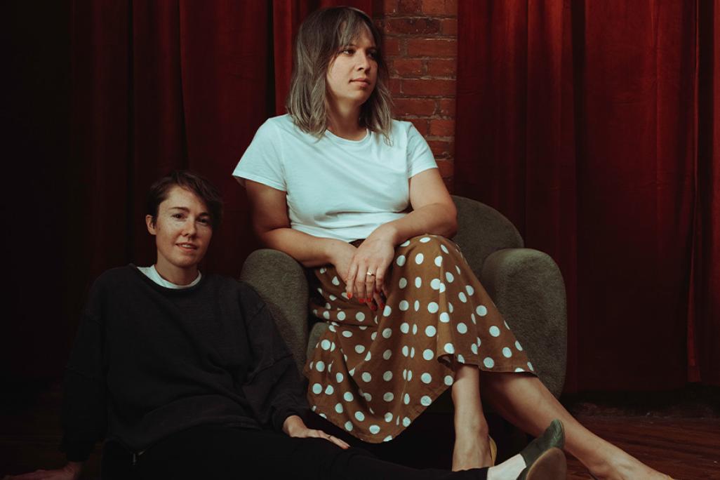 Andrew Yee and Caroline Shaw pose together in front of 2 red velvet curtains and a strip of brick wall. Shaw sits on the floor to the left of Yee, smiling slight at the camera and wearing a tee shirt layered underneath a grey sweater and black pants. Yee sits in a chair next to Shaw, wearing a white tee and a brown skirt with white polka dots.