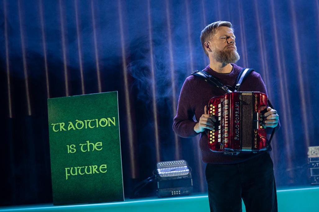 Man performs accordion on stage in front of a sign that reads 'Tradition is the Future'