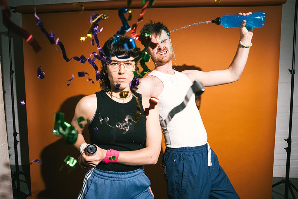 Two energetic performers in athletic wear strike playful poses against an orange backdrop, spraying water and launching colorful confetti streamers toward the camera.