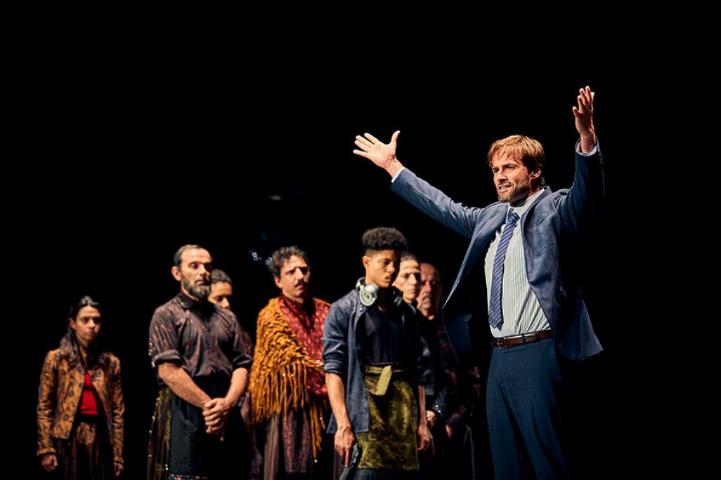 A performer on stage poses with outstretched arms and cast members in the foreground. 