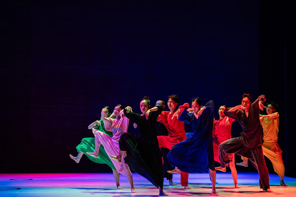 Members of Cloud Gate march across a rainbow lit stage wearing colorful gowns. They move in unison with one leg raised, mid large step.