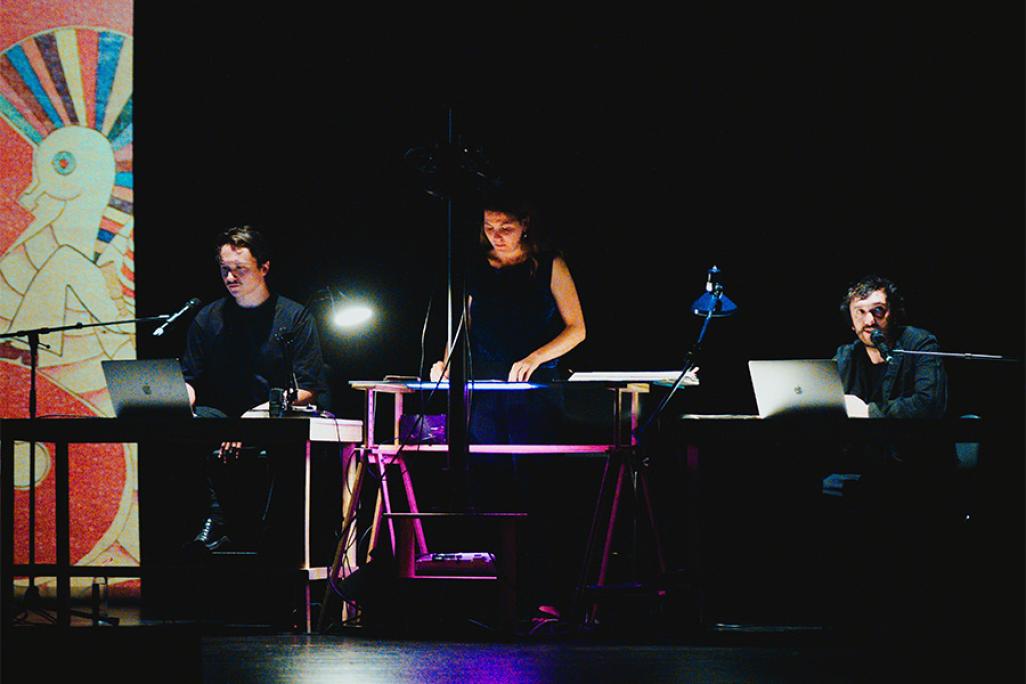 Cuervo performers seated and standing at illuminated workstations with laptops and microphones during a multimedia live performance on a darkened stage.
