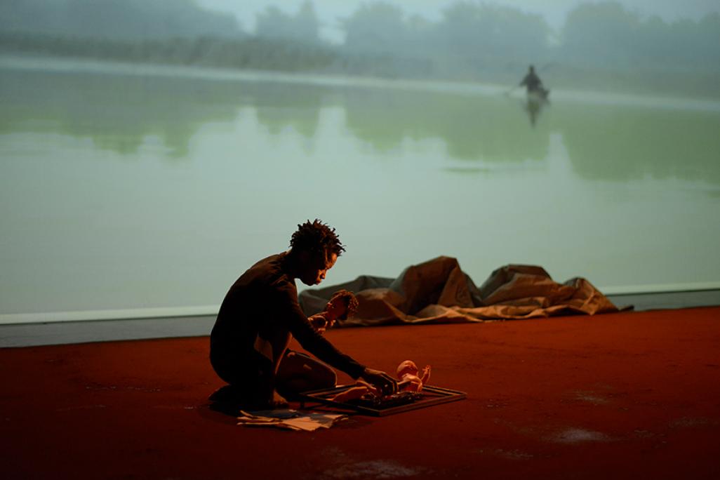 Faustin Linyekula crouches on stage with image of body of water projected behind him