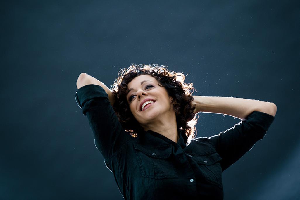 Portrait of luciana with curly hair smiling and leaning back with her hands behind her head, illuminated by soft backlighting against a dark, moody background