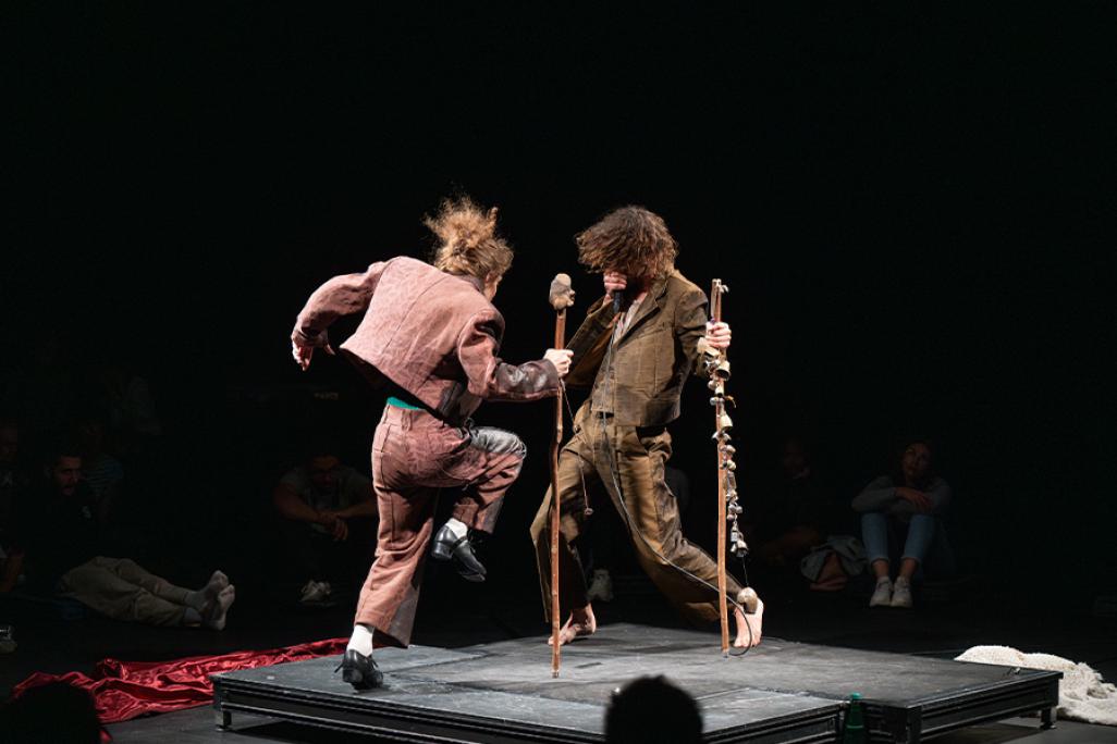 Performance image from Mirlitons featuring Hainaux and Chaignaud performing in the center of a theater on a small square stage. Silhouettes of audience members are seen in the foreground and background. Chaignaud faces away from the camera as he lifts on leg in the air, tap dancing. He wears pink pants and a jacket, holding a large wooden stick. Hainaux faces towards the camera and Chaignaud, wearing a brown suit and holding a wooden stick with bells attached to it. He beatboxes into a microphone.