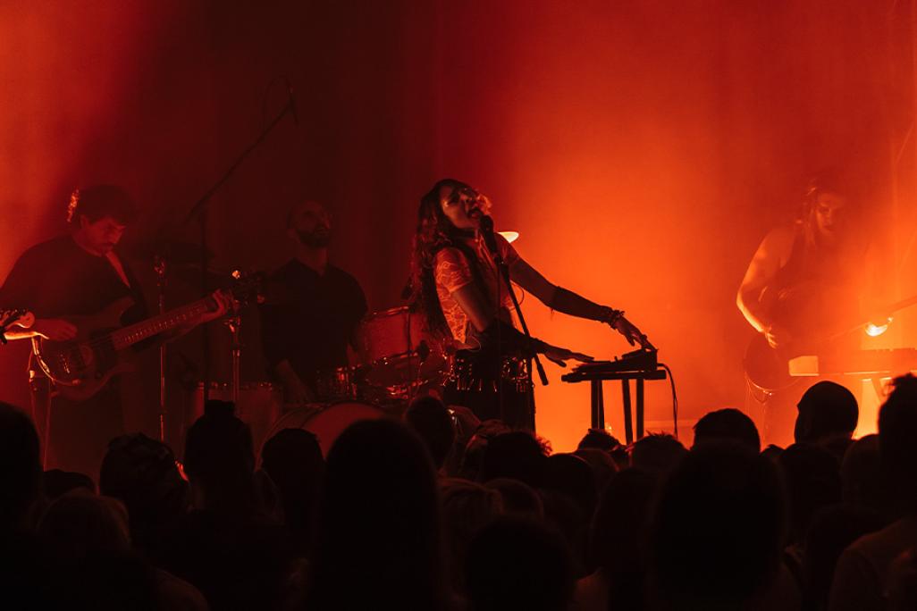SPELLLING performs on stage, lit by orange light. There are the silhouettes of audience members in the foreground and blurred silhouettes of SPELLLING's band behind them performing on stage.