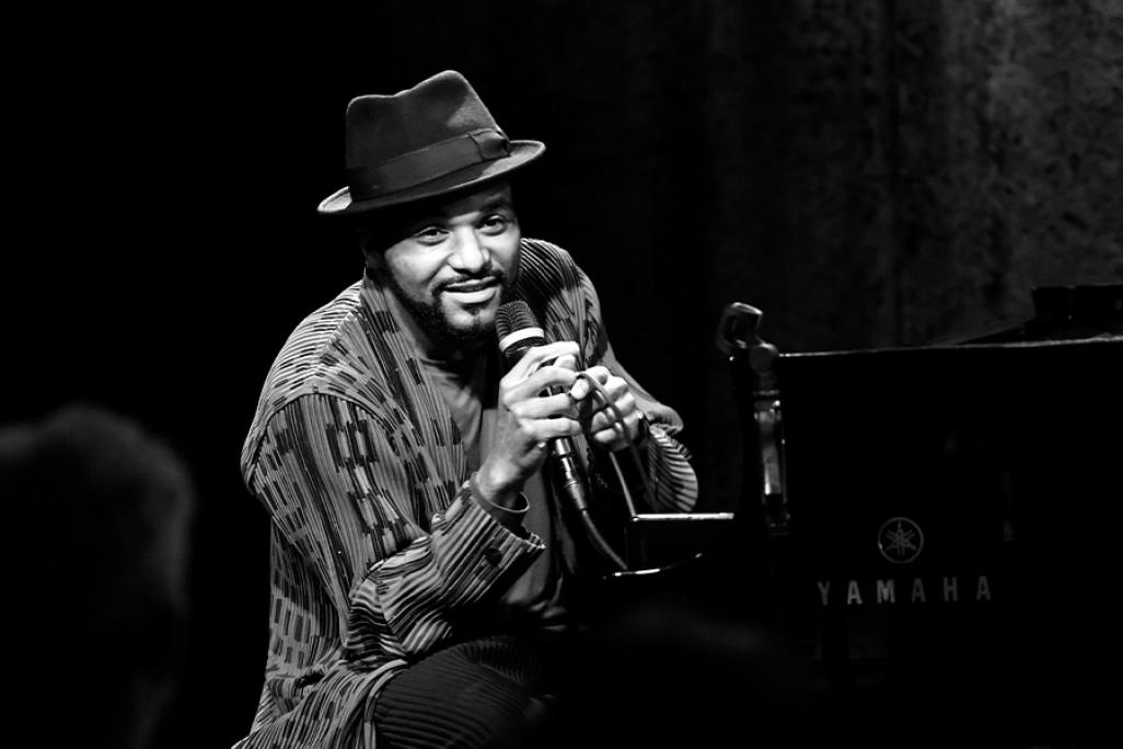 Black and white image of Sullivan Fortner speaking into a mic while sitting at a piano