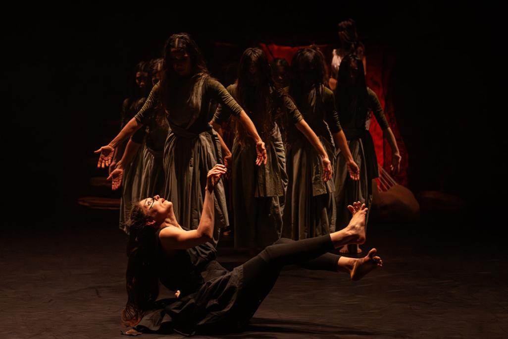 A group of dancers stand with arms partially stretched out to their sides on a dark stage wearing grey dresses. They look downwards at another dancer, wearing all black, seated on the floor with her arms and legs extended up and outward, looking startled.