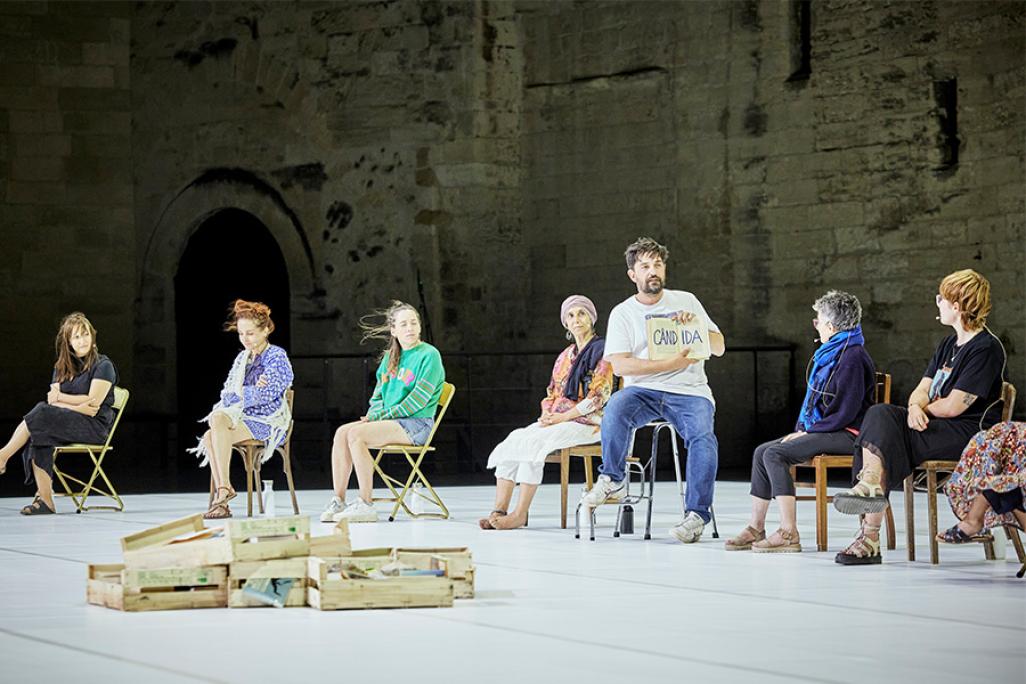 A group of people sit on stage in chairs with a pile of cabinet drawers in front of them on the floor. One of the participants holds open a book.
