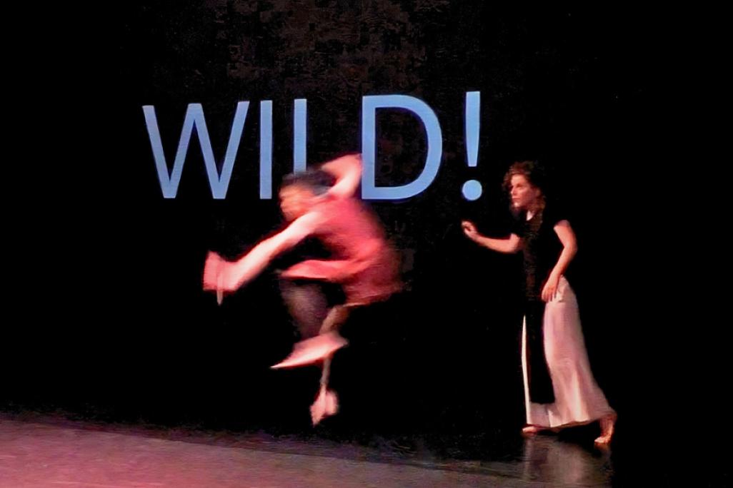 Two dancers perform in front of black back drop with the world 'WILD!' projected onto it. One of the dancers wears a red shirt and jumps mid air while the other performer steps towards them with one arm outward.