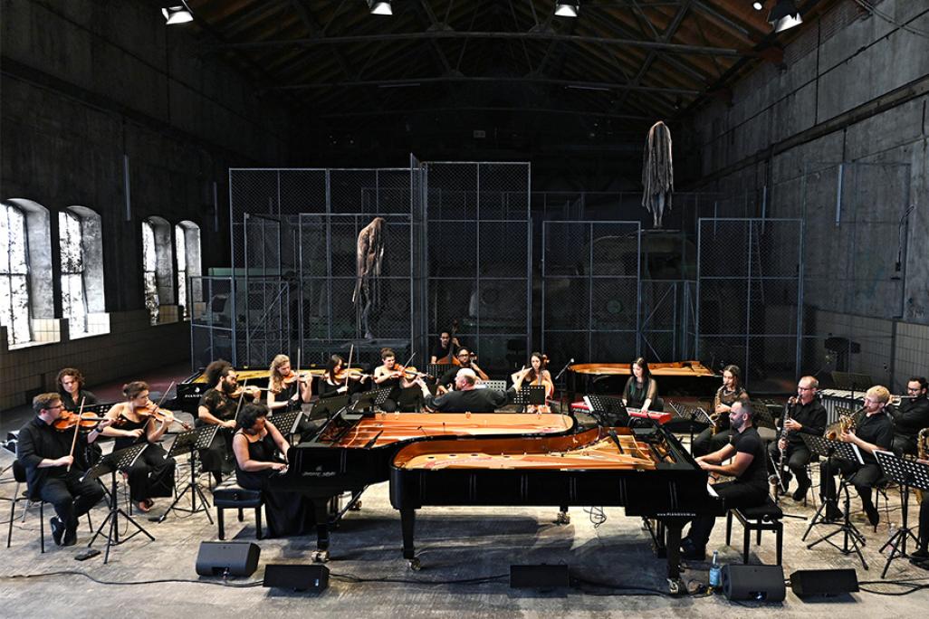 Musicians perform at two pianos, surrounded by a large orchestra, in a warehouse like space