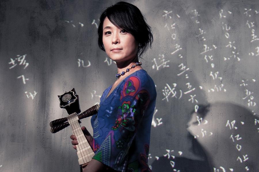 Wu Man stands with a pipa in front of a wall with writing
