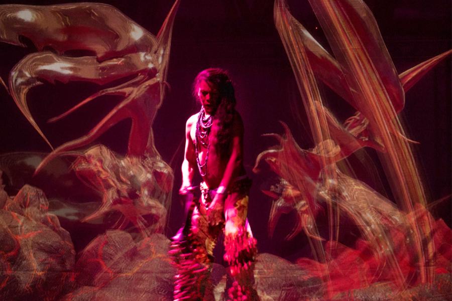Performer on stage lit by a pink light with abstract sculptures surrounding them
