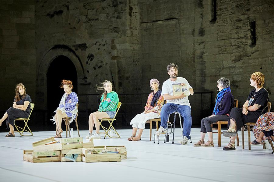 A group of people sit on stage in chairs with a pile of cabinet drawers in front of them on the floor. One of the participants holds open a book.