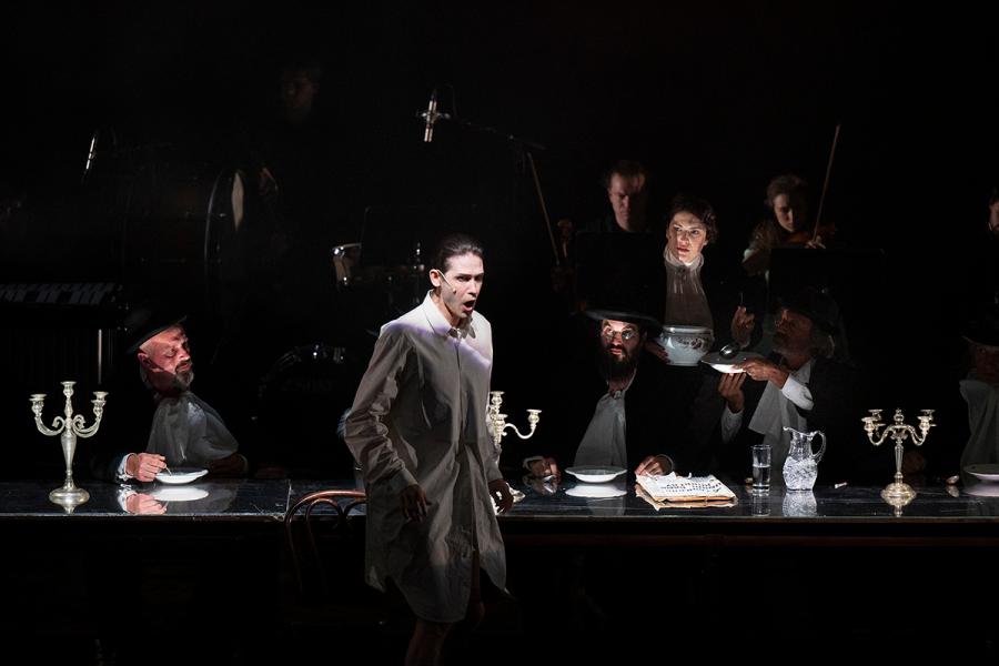 A central performer stands illuminated at a long dinner table surrounded by seated figures in dark formal attire, with candlelight, musicians, and shadowed staging in "Kafka's Letter to His Father"