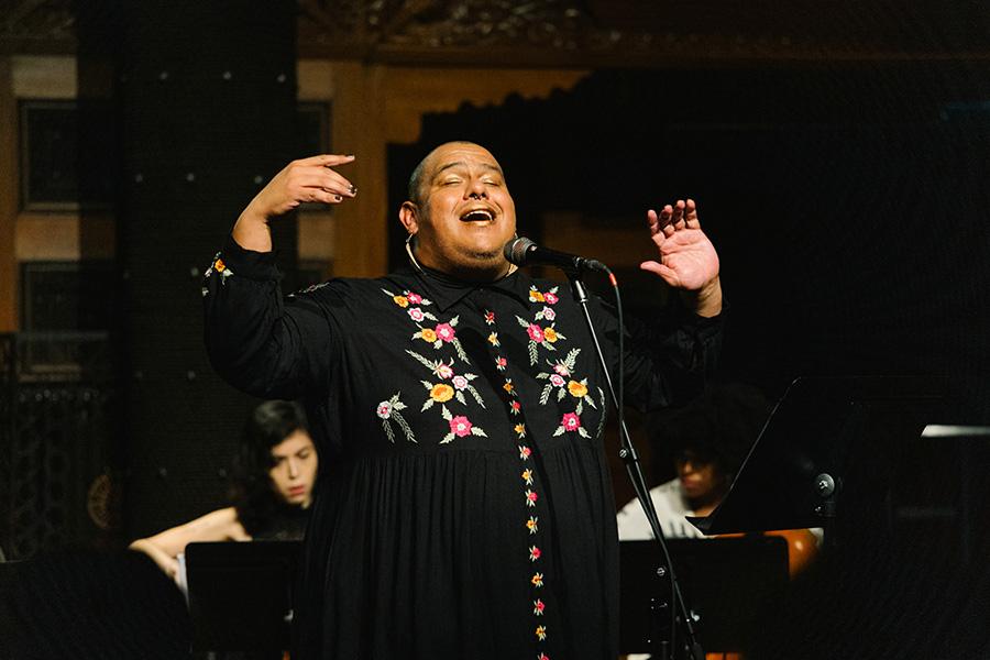 Dorian Wood performs on stage, lit by warm light. They wear a floral embroidered black dress and lift their hands in the air as they sing into a microphone. Two musicians sit behind them, playing instruments partially obstructed by music stands.
