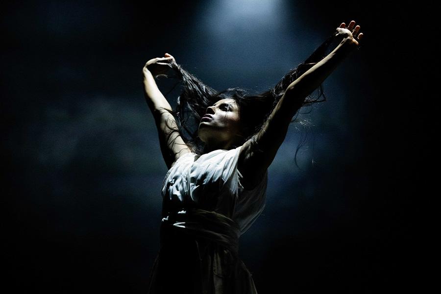A dancer poses with arms raised above their head on a stage with a dark backdrop and smoke floating in the air.